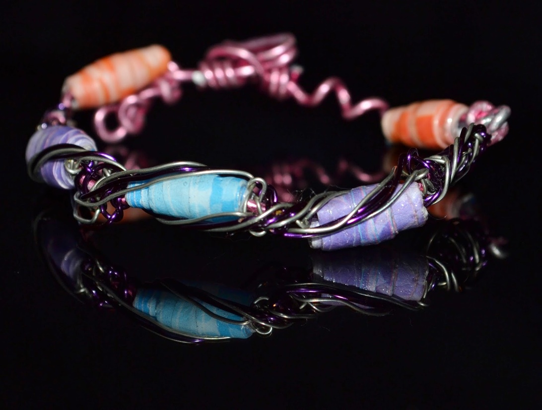 Ett vackert handgjort armband med återbrukade pärlor i blå, lila och orange toner, sammanfogade med rosa och lila metalltråd mot en svart bakgrund.